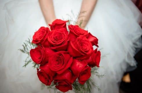 A bride in a white dress holding a large bouquet of bright red roses with a bit of greenery
