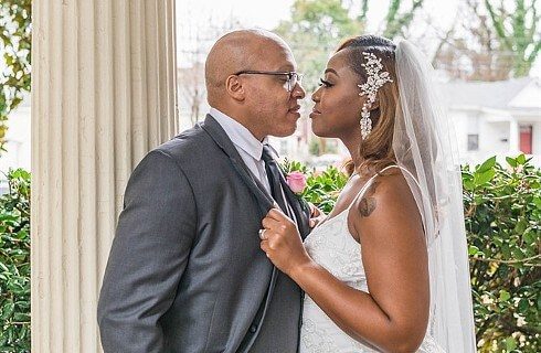 An African American bride and groom standing facing each other outdoors near a white column