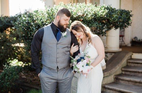 A bride putting her head on the shoulder of a groom at the base of stone steps outside a home