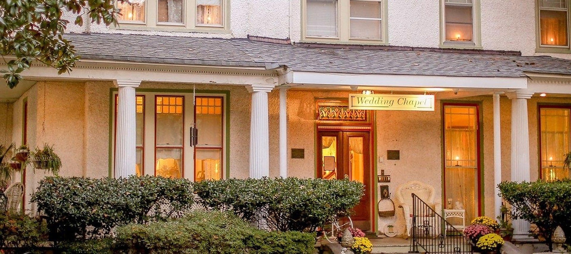 A beautiful white home with a wrap around porch, white columns, golden lights at night and bushes