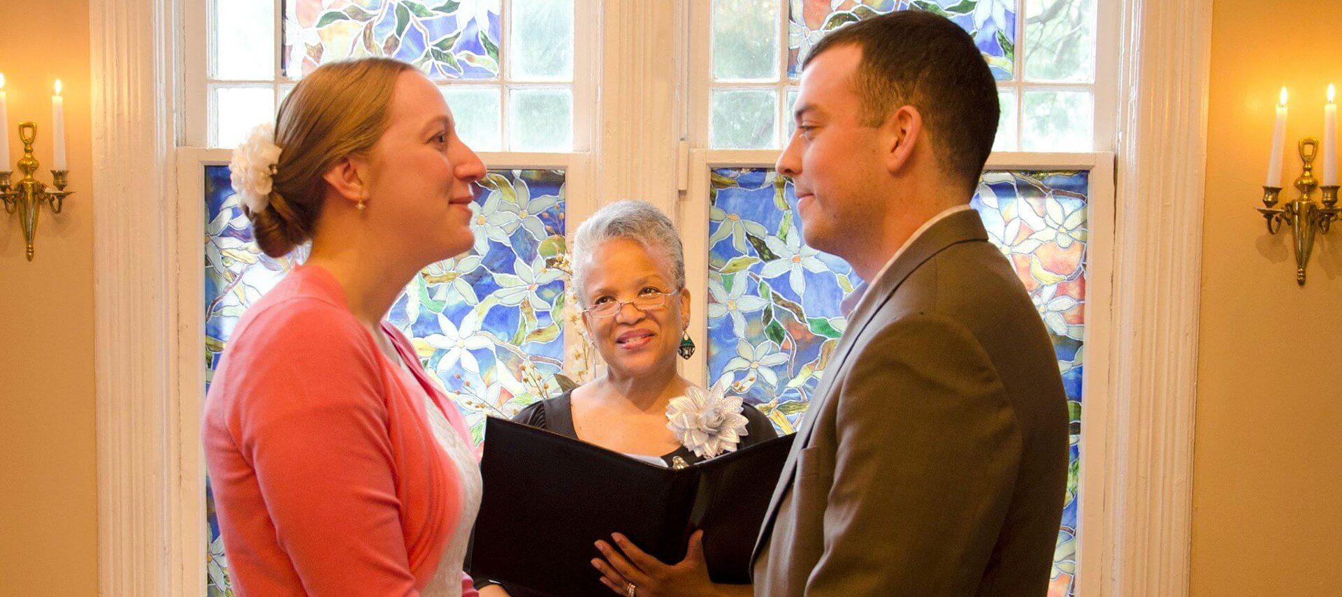 A bride and groom standing in front of a woman officiating their wedding in a room with stained glass windows