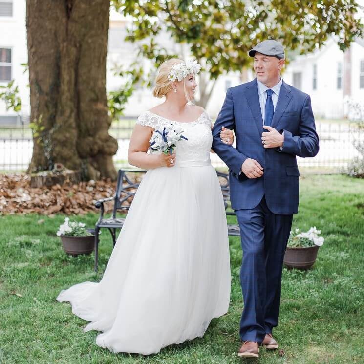 A bride with blonde hair and a groom in a tux and cap walking outdoors in a grassy area by a large tree