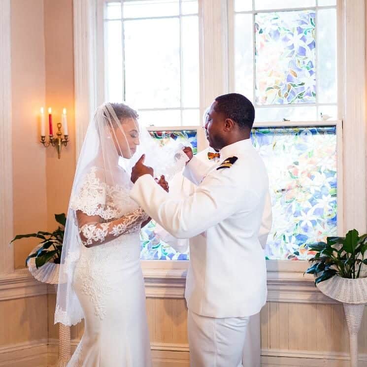 An African American couple standing by a large stained glass window as the groom lifts the bride's veil