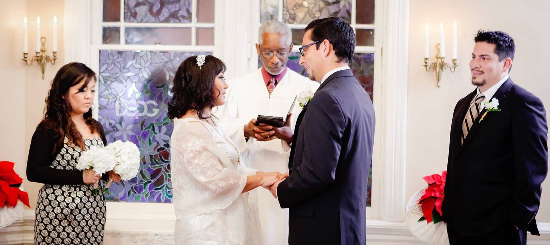 A bride and groom and their attendants stand at an alter with an officiant in front of a stained glass window