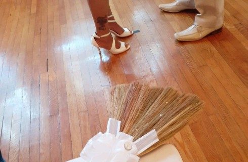 A woman's feet in high heels next to a man's feet in dress shoes standing near a broom with a white ribbon