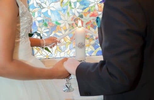 A bride and a groom standing holding hands and lighting a candle together in front of a stained glass window