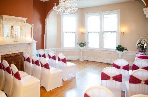 A small indoor chapel with bright windows, a fireplace with mantel and white chairs with red ribbons