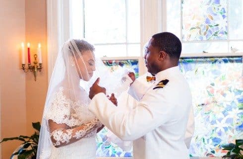An African American couple standing by a large stained glass window as the groom lifts the bride's veil