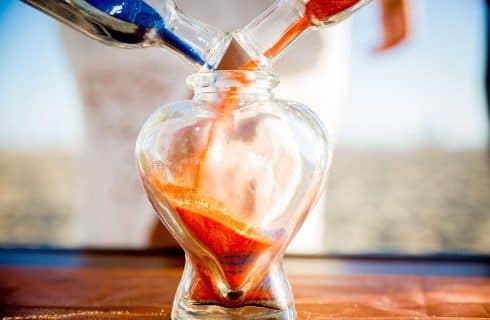Two bottles of sand, red and blue, being poured into a larger bottle in the shape of a heart